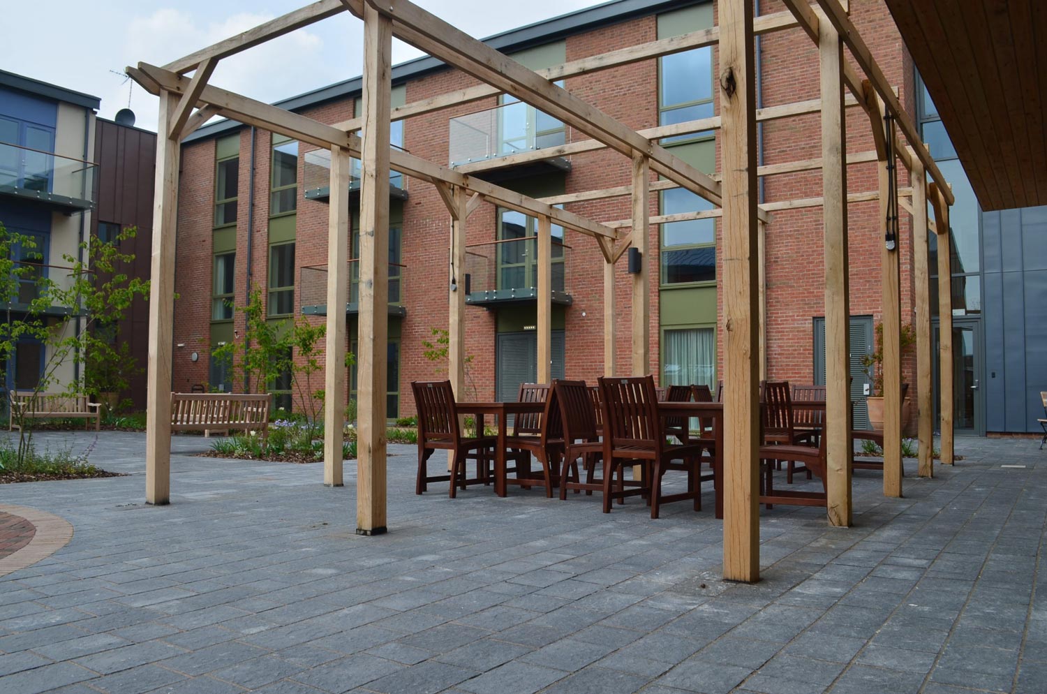 A wooden pergola with tables and chairs sits on a paved courtyard area in front of a brick building.
