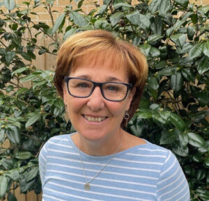 Michelle Moorcraft, wearing a light blue and white striped shirt and glasses, smiles in front of a green bush.