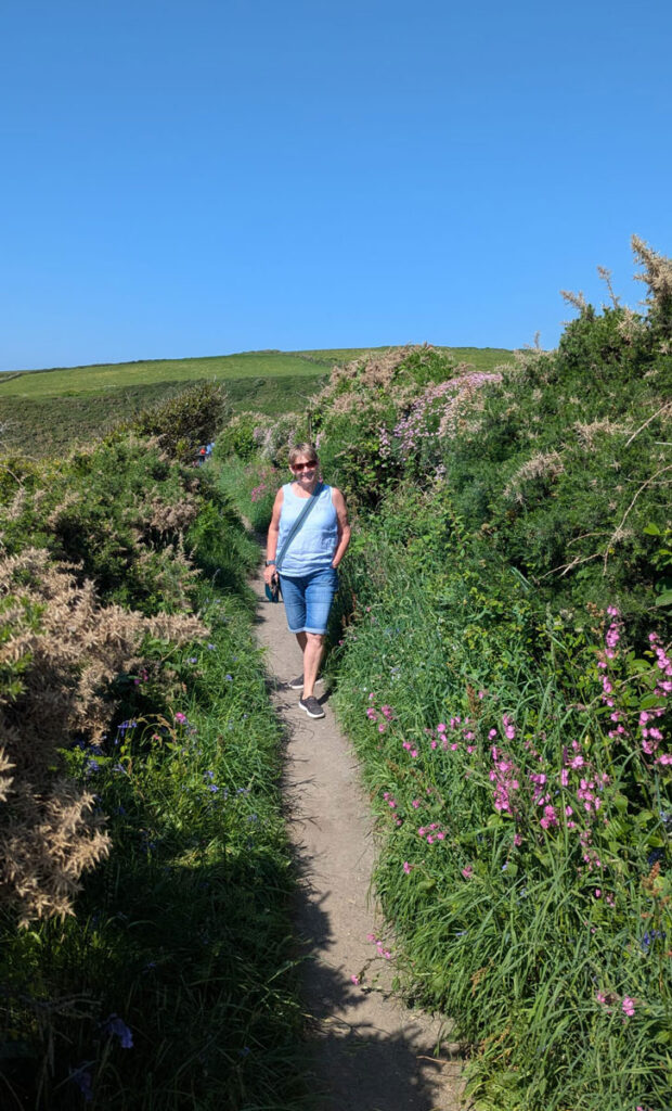 Lil Bowers walking along a narrow countryside path bordered by tall grass and wildflowers under a clear blue sky.