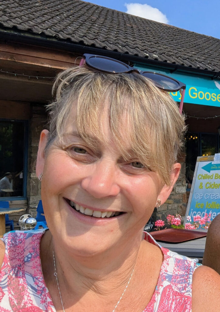 Close-up portrait of Lil Bowers smiling outdoors, with short hair and sunglasses resting on her head, taken outside a café on a bright day.