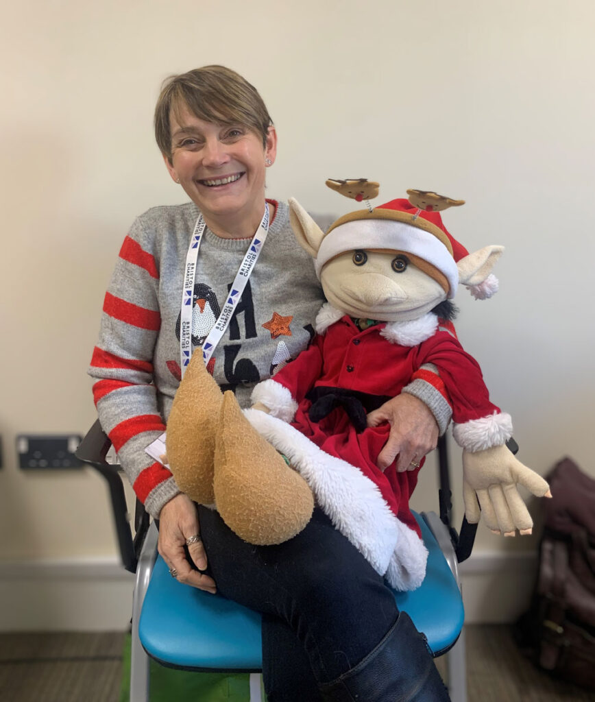 Lil Bowers smiling while seated indoors, holding a large festive elf puppet dressed in a red Christmas outfit.