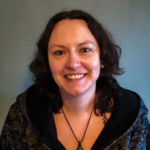 Beth Cox Smith, wearing a patterned dark jacket and a necklace with a green pendant, smiles against a light blue-gray background.