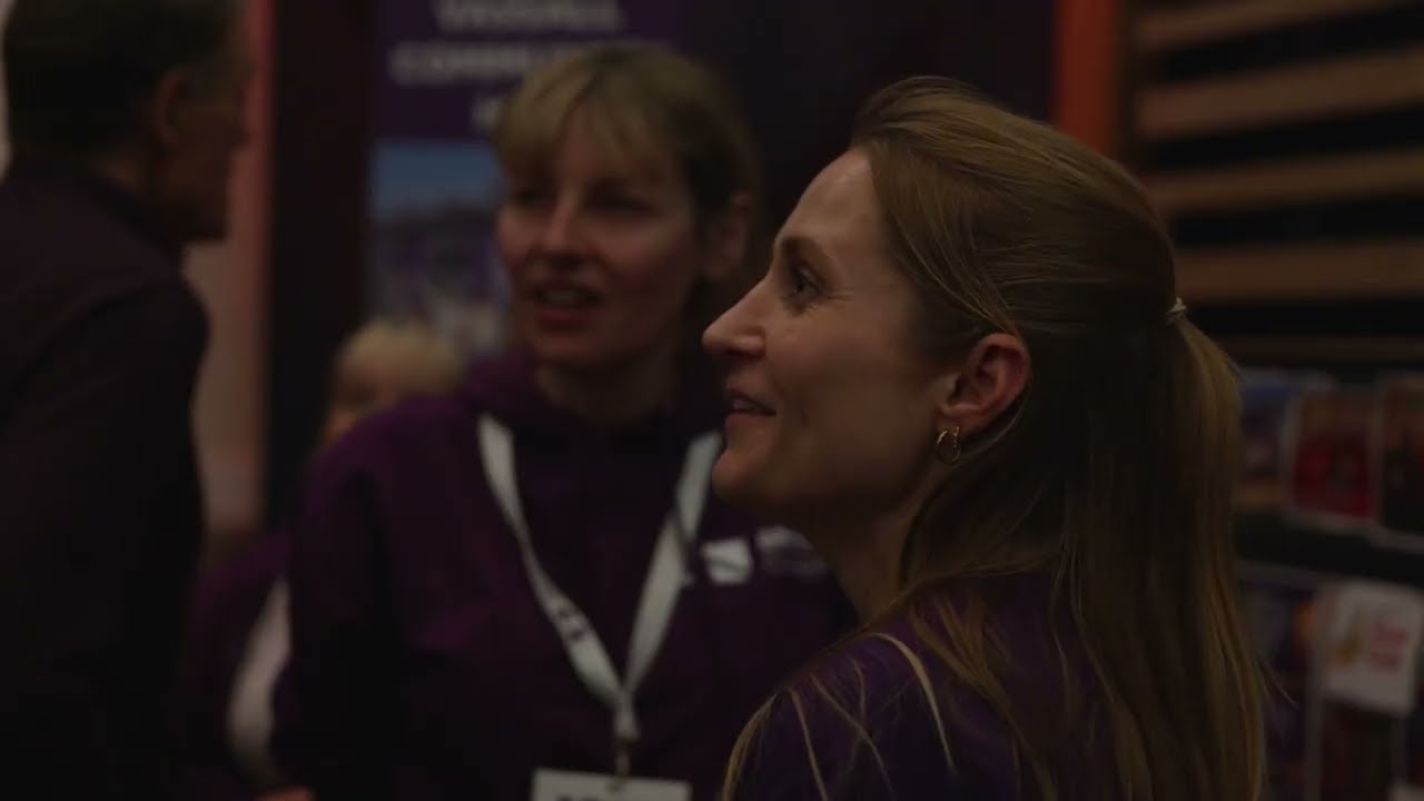 A woman with light brown hair pulled back in a ponytail wears a purple top and gold hoop earrings, smiling slightly as she looks to the right, with two blurred figures in purple clothing behind her.