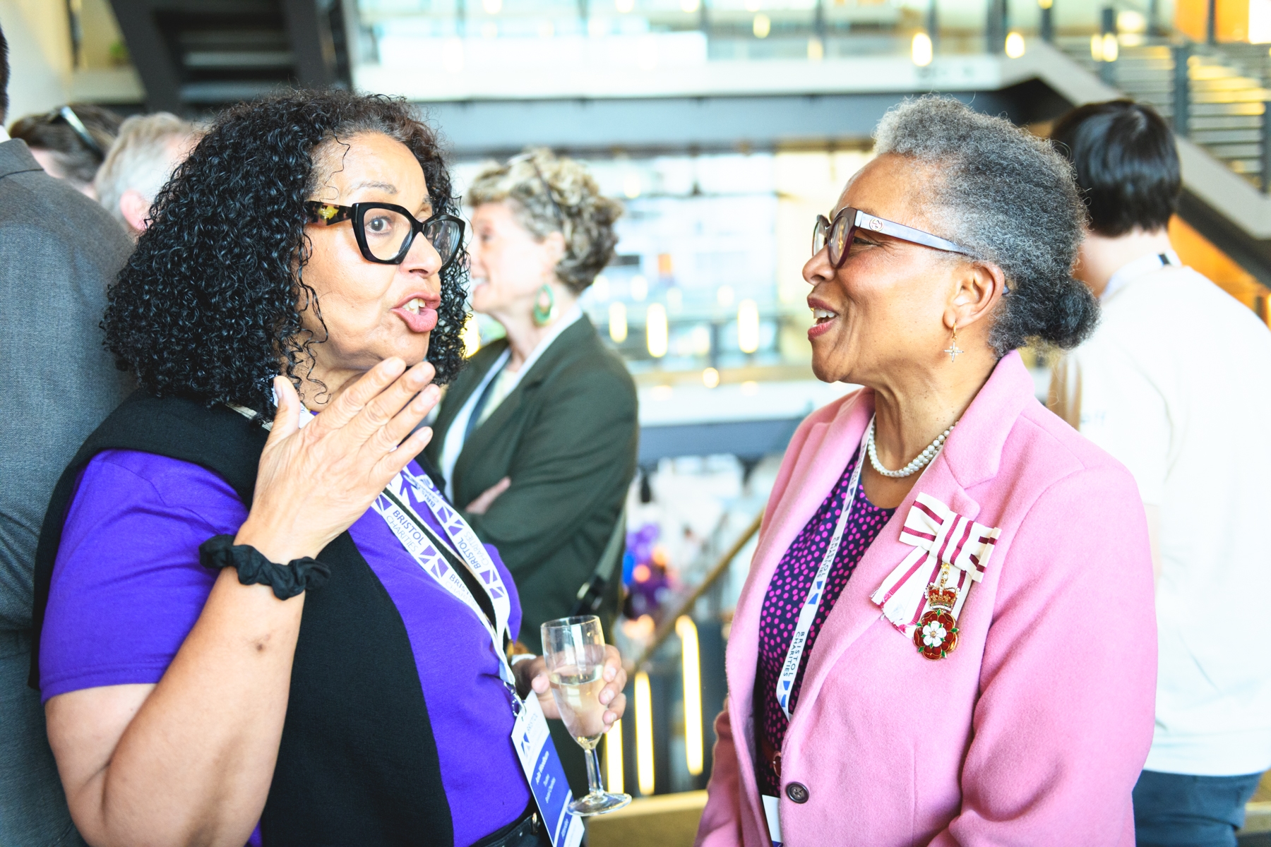 Two women, one in a purple shirt and dark vest, the other in a pink jacket and patterned top, converse at a Bristol Charities event.