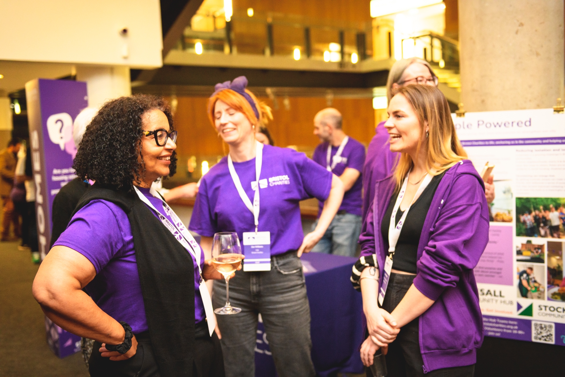 Three women in purple shirts converse at a Bristol Charities event.
