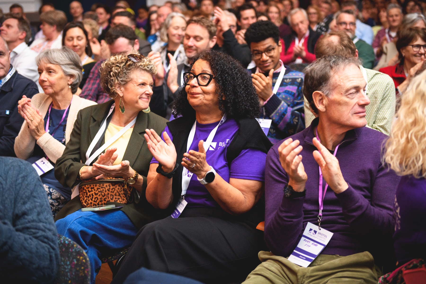 Anita, Rachel and Andy sit in a crowd, clapping their hands and wearing lanyards with badges.